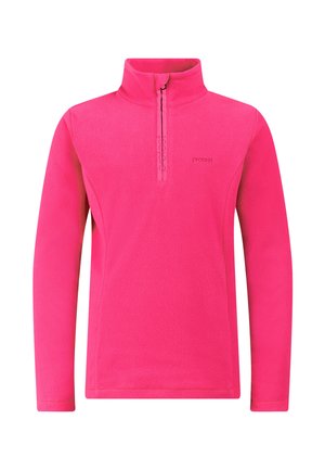 Jonge vrouw die een roze fleece draagt, paarse broek, zwarte laarzen en een roze-beige gebreide muts, staand tegen een effen lichte achtergrond.