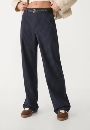 Personne portant un pantalon large bleu marine à fines rayures avec ceinture noire, une veste beige courte, un haut blanc et des baskets marron, debout sur un sol blanc.