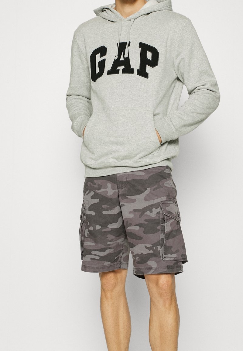 Personne portant un sweat à capuche gris clair avec le texte "GAP" et un short cargo camouflage gris avec des poches, debout les mains dans les poches du sweat à capuche.