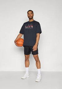 Černé oversized tričko s červeným grafickým nápisem, v kombinaci s černými šortkami a bílými teniskami. Model drží oranžový basketbalový míč.