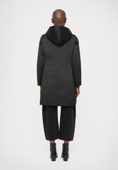 Cappotto imbottito nero con cappuccio, design lungo fino al ginocchio e maniche lunghe. Indossato sopra pantaloni neri a gamba larga e stivali neri con tacco.