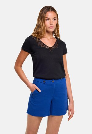 Mujer con una camiseta negra de manga corta con escote de encaje y pantalones cortos azul brillante con botones, de pie con una mano en el bolsillo.