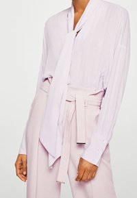 Licht paarse blouse met een strik-kraag, lange mouwen en knoopmanchetten, gecombineerd met bleekroze broek met een ceintuur in de taille.