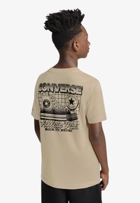 Μπεζ βαμβακερό μπλουζάκι με μαύρο γραφικό ενός αθλητικού παπουτσιού Converse και κείμενο "CONVERSE Athletic Dept." στην πλάτη. Κοντά μανίκια.