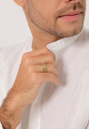 Gold ring with a textured, braided design worn on a hand, partially visible beside a white, collared shirt. Skin tone appears light.