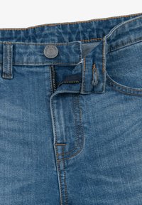 Jeans denim w średnim praniu z kontrastującymi brązowymi szwami, z suwakiem, zapięciem na guzik i widocznie przeszytymi kieszeniami.