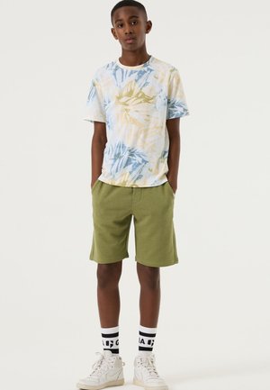 Lichtblauw, geel en groen patroon T-shirt met korte mouwen, gecombineerd met olijfgroene korte broek. Geaccentueerd met witte sneakers en zwarte sokken.