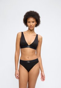 Γυναίκα με σγουρά μαλλιά που φοράει μαύρο bralette Calvin Klein και ταιριαστά σλιπ με ψηλή μέση, στέκεται μπροστά σε ένα απλό φωτεινό φόντο.