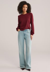 Bordo teksturirana bluza dugih rukava uparena s svijetloplavim trapericama širokih nogavica i smeđim cipelama špic, s visokim strukom i suptilnim varijacijama u pranju.