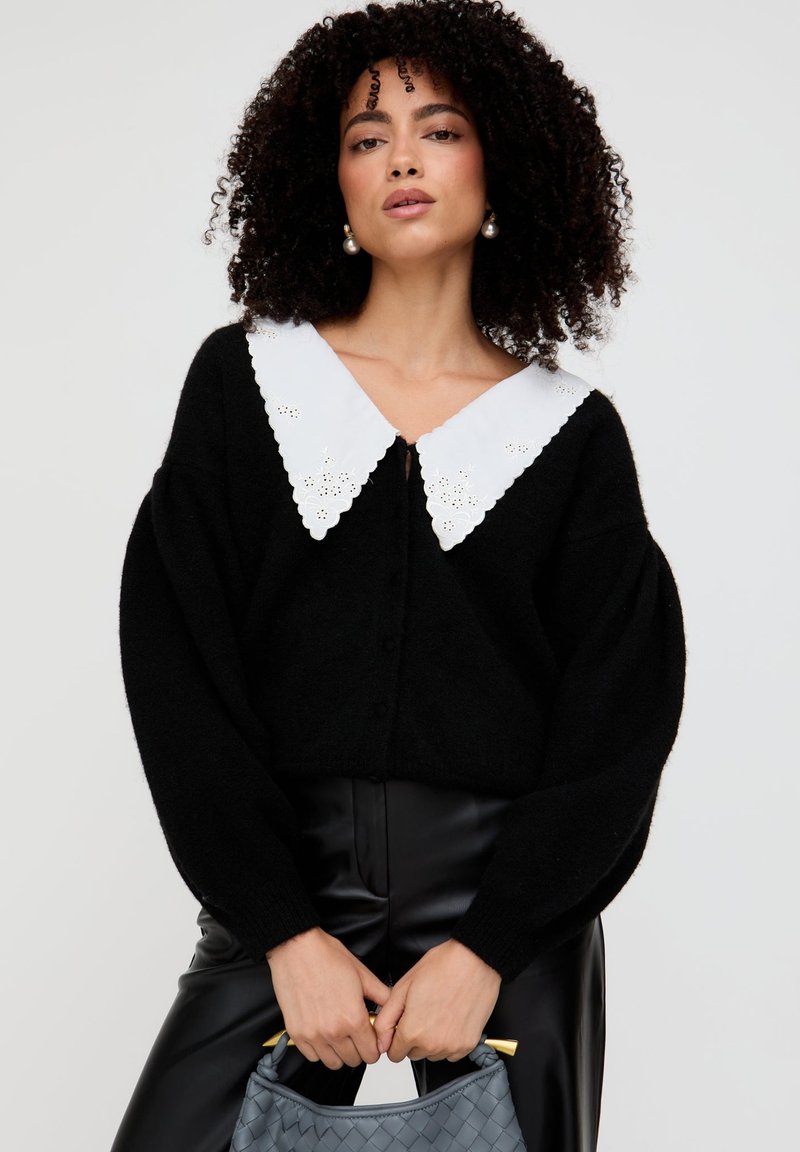 Schwarze gestrickte Strickjacke mit großem weißem, gewelltem Kragen mit floralen Ausschnitten. Knöpfe vorne; das Model hält eine graue Tasche.