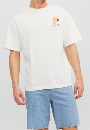 Mann trägt weißes Santorini-Grafik-T-Shirt und hellblaue Jeans-Shorts vor einfarbigem Hintergrund.