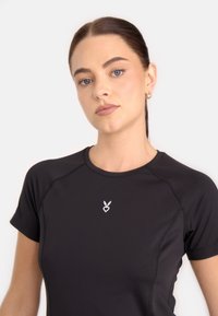 Melnā sporta T-kreklu ar īsām piedurknēm, ar baltu logo uz krūšu daļas, gluda tekstūra un pieguļošs dizains.