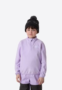 Polarowy sweter w jasnym lawendowym kolorze z kołnierzem na zamek, w połączeniu z ciemną, wzorzystą dzianą czapką. Dziecko nosi spodnie narciarskie w dopasowanych kolorach.