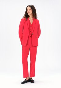 Completo rosso su misura con un blazer con rever a punta, un gilet aderente con bottoni e pantaloni slim. Indossato con mocassini neri.