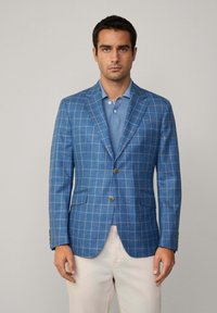 Blazer a quadri blu realizzato in tessuto intrecciato, con rever a punta, due tasche frontali e bottoni marroni. Indossato sopra una polo azzurra chiara.