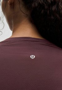 Bordová atletická košile s texturovaným povrchem, kulatým výstřihem a malým bílým logem poblíž krku. Má přiléhavý design a šité detaily.