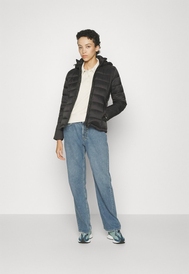 Chaqueta acolchada negra con líneas de acolchado horizontales, capucha y bolsillos frontales con cremallera. Combinada con una polo color crema y jeans de color azul claro.