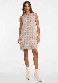 Mouwloze jurk van een lichtroze geweven stof met een ruitpatroon. Heeft franjes aan de zoom en is gecombineerd met beige enkellaarsjes.