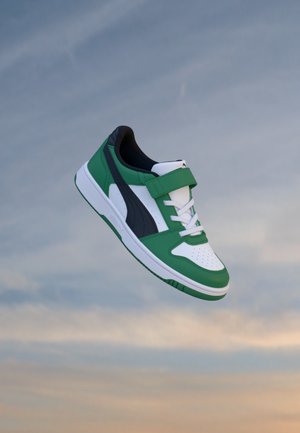Baskets vertes, blanches et noires avec lacets et bande velcro flottant sur un fond de ciel au coucher du soleil.