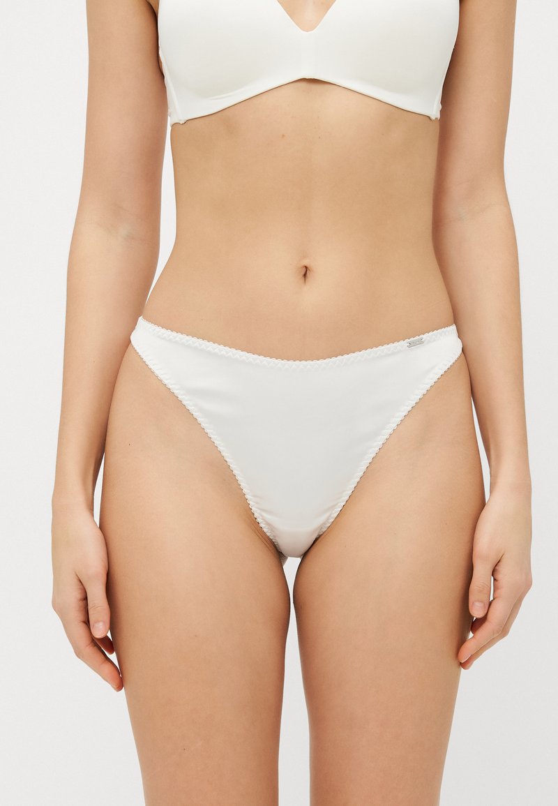 Perizoma bianco in satin con una texture liscia, caratterizzato da delicati bordi ondulati e un piccolo tag con logo sul lato. Design minimalista con vita bassa.