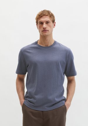 Jonge man met kort blond haar die een effen blauw T-shirt met korte mouwen en een bruine broek draagt, staand met handen in de zakken.