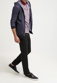 Felpa con zip di colore navy indossata sopra una camicia colletto bordeaux, abbinata a pantaloni neri aderenti e sneakers nere con dettagli testurizzati.