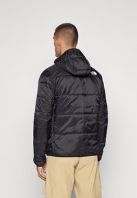 Mann med ryggen til iført en svart boblejakke med hette og beige shorts mot en ensfarget lys bakgrunn.