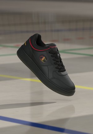 Baskets noires avec lacets noirs, logo jaune et rouge sur le côté, bordure rouge et logo Champion sur la languette et le talon, flottant au-dessus du sol d’une salle de sport.
