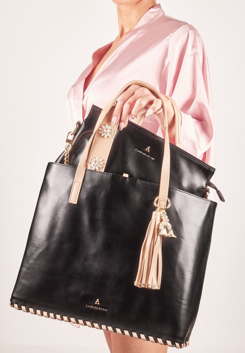 Sac en cuir noir avec des accents taupe, featuring des embellissements floraux, un pompon et des coutures le long du bord inférieur.