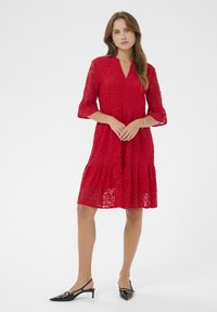 Rochie roșie din dantelă cu mâneci tip clopot 3/4, decolteu în V, tiv evazat și model floral texturat; purtată cu tocuri slingback negre.