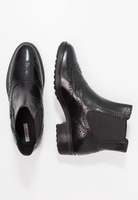 Botas de tobillo de cuero negro con detalles de brogue, paneles laterales elásticos y suela de goma. Presenta un tacón bajo en bloque y textura suave.