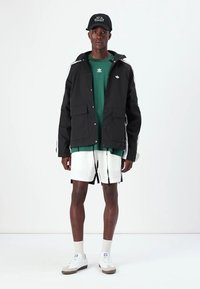 Casaco impermeável preto com riscas brancas, sweatshirt verde, calções brancos com pormenores pretos e sapatilhas cinzentas, usados com uma capa preta.