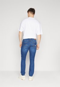 Blue denim jeans dengan potongan slim, saku belakang polos, dan patch kulit coklat di pinggang. Dipadukan dengan kaos lengan pendek putih.