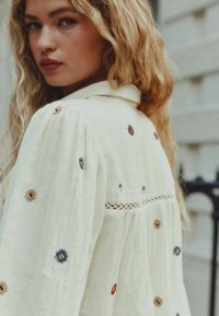White embroidered blouse featuring intricate diamond patterns in blue, brown, and red. Soft, textured fabric with a classic collar and lace detail.
