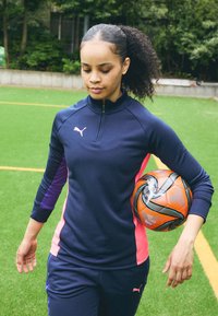 Námornícky zipsový športový top s ružovými a fialovými akcentmi, v kombinácii s námorníckymi nohavicami. Drží oranžovú a čiernu futbalovú loptu, umiestnený na zelenej tráve.
