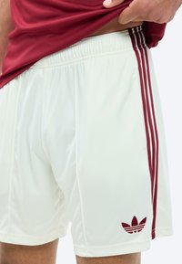 Shorts de sport blancs avec logo Adidas bordeaux et trois bandes sur le côté, portés par une personne levant un t-shirt bordeaux.