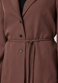 Bruine riemjas met zwarte knopen, gedragen over een zwarte top, die details van de middelste sectie en de stoftextuur toont.