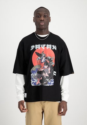 Giovane uomo con una maglietta nera con grafica, testo giapponese, un cerchio rosso e una scena illustrata sopra una camicia bianca a maniche lunghe e pantaloni beige.