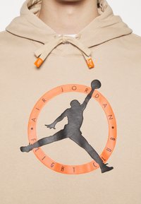 Sudadera con capucha beige con cordón y detalles en naranja. Presenta una silueta negra de una figura saltando dentro de un audaz logo circular naranja.