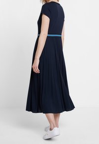 Marinblå klänning med veck och korta ärmar, kompletterad med ett blått accentbälte. Golvlång design med en slät textur, kombinerat med vita sneakers.