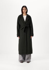 Μακριά σκούρα πράσινη καπαρντίνα από χοντρό ύφασμα με διπλό κούμπωμα, μεγάλα πέτα και μέση με ζώνη, συνδυασμένη με φαρδιά γκρι παντελόνια.