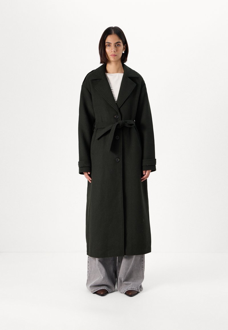 Μακριά σκούρα πράσινη καπαρντίνα από χοντρό ύφασμα με διπλό κούμπωμα, μεγάλα πέτα και μέση με ζώνη, συνδυασμένη με φαρδιά γκρι παντελόνια.