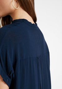 Blusa blu navy con una texture morbida, maniche corte arrotolate e un design fluido. Presenta una piega sul retro per un maggiore movimento.
