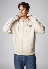 Cremefarbener Kapuzenpullover mit Reißverschluss und Kängurutasche, schwarzer Schriftzug "EXPECT NOTHING" auf der Brust, mit Kordelzug und gerippten Bündchen.