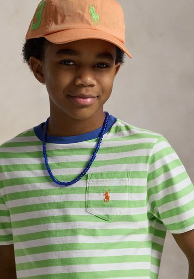 Chlapec nosi oranžovú čiapku, zeleno-biela pruhovanú košeľu s predným vreckom a modrý náhrdelník z korálikov na neutrálnej pozadí.