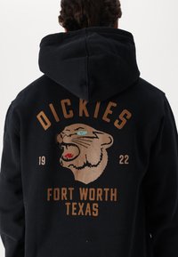 Sudadera negra con un gran gráfico de un pantera rugiente en beige y rojo, con "DICKIES" y "FORT WORTH TEXAS" en letras gruesas y marrones.