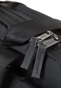 Dettaglio di uno zaino in nylon nero con zip doppie e tiranti in pelle, e una finitura testurizzata. Gli accenti includono cuciture resistenti e spallacci regolabili.