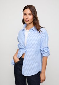 Svijetloplava košulja s dugim rukavima i gumbima, bijelim vertikalnim prugama, s džepom na prsima i podvrnutim rukavima, u kombinaciji s tamnim trapericama.