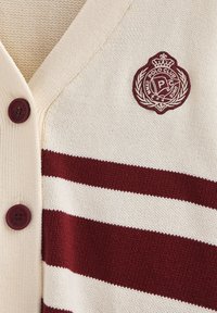 Krämfärgad cardigan med vinröda horisontella ränder, texturerat stickat tyg, två vinröda knappar och en rund broderad logotyp med krona.