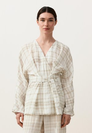 Kvinde iført lysebeige plaid slå-om jakke med vide ærmer og matchende bukser, stående mod en ensfarvet baggrund.